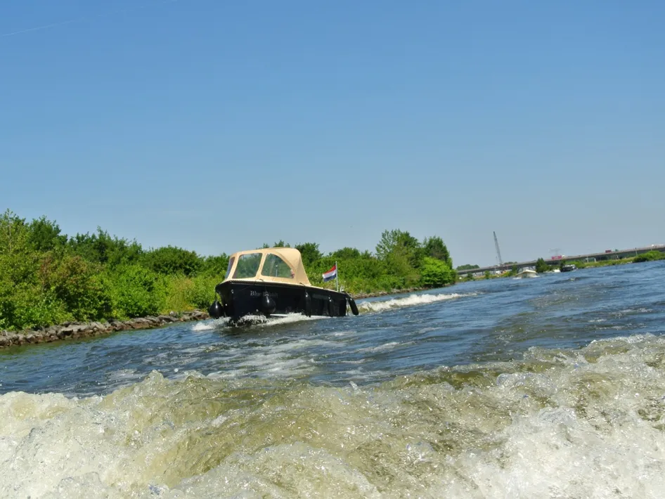 Die Schaluppe Blue Dolphin fährt auf dem Fluss davon, vom Wasserspiegel aus gesehen, mit grünem Ufer und einer Brücke im Hintergrund.