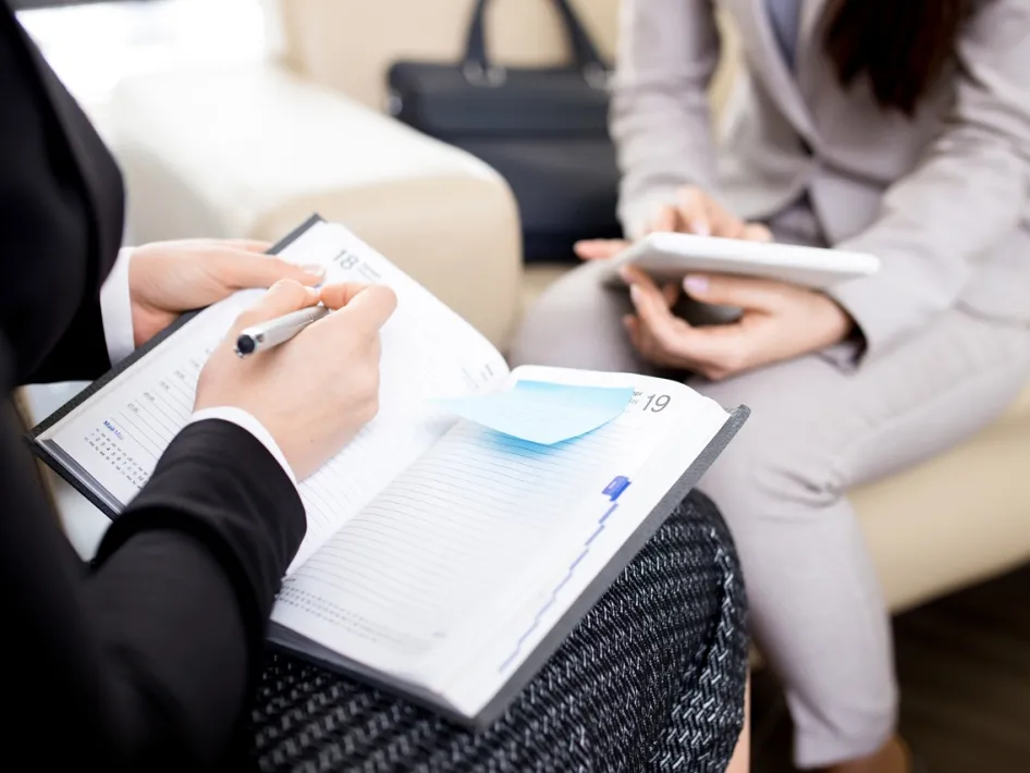 Twee vrouwen in gesprek tijdens een professioneel Spreekuur, met notitieboek en tablet in de hand.
