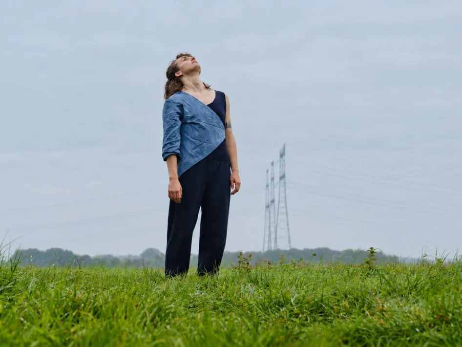 Sarah Soethoudt staat stil in het landschap tijdens Stiltewandeling AARDE(N), met gras en een open lucht op de achtergrond.