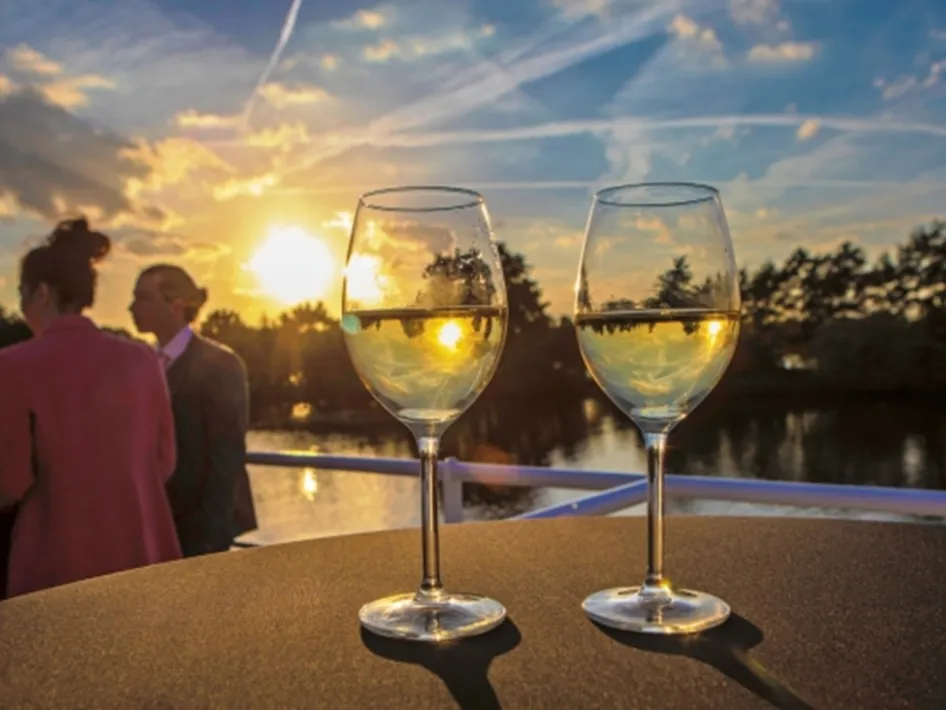 Twee glazen witte wijn op het dek tijdens de Sunset Cruise, met een prachtige zonsondergang boven de Maas op de achtergrond.