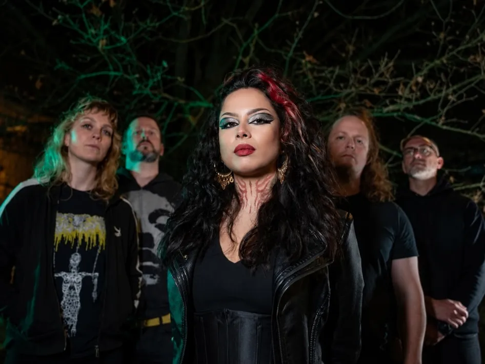 De metalband The Fifth Alliance poseert 's avonds in het bos, met zangeres op de voorgrond en de bandleden daarachter.