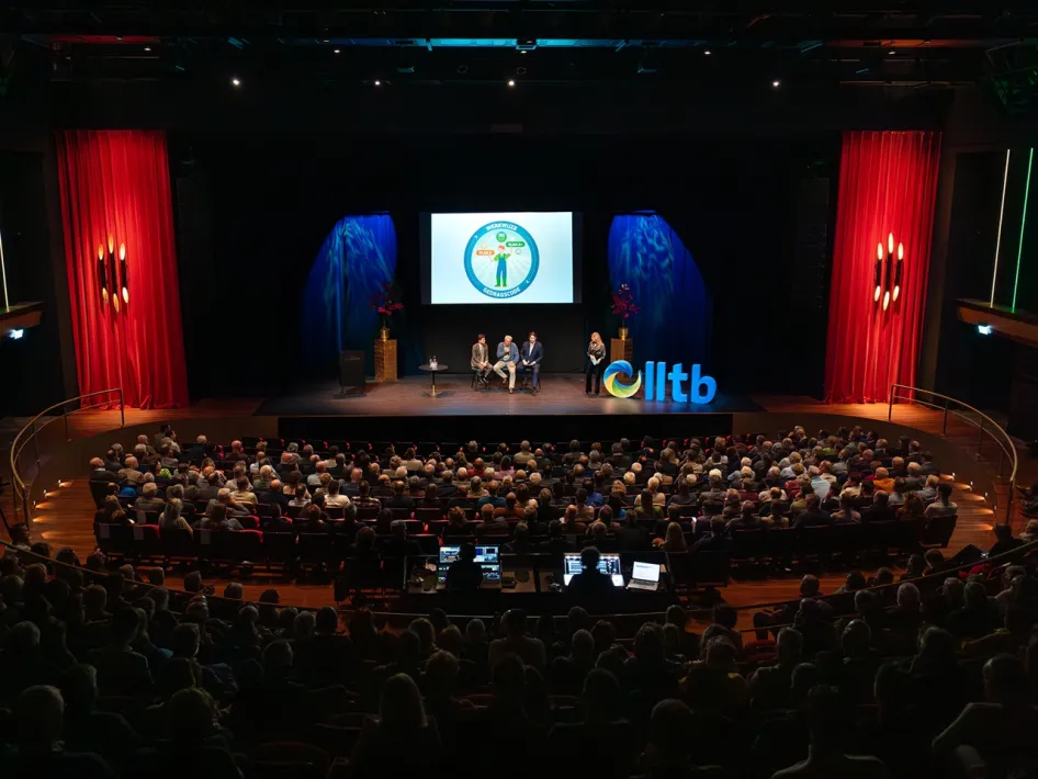 Drukke theaterzaal tijdens een lezing of congres met publiek en sprekers op het podium.