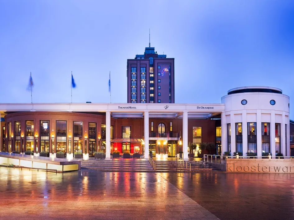 Frontansicht des TheaterHotel De Oranjerie am Abend, mit beleuchteter Fassade und einem verregneten Platz im Vordergrund.