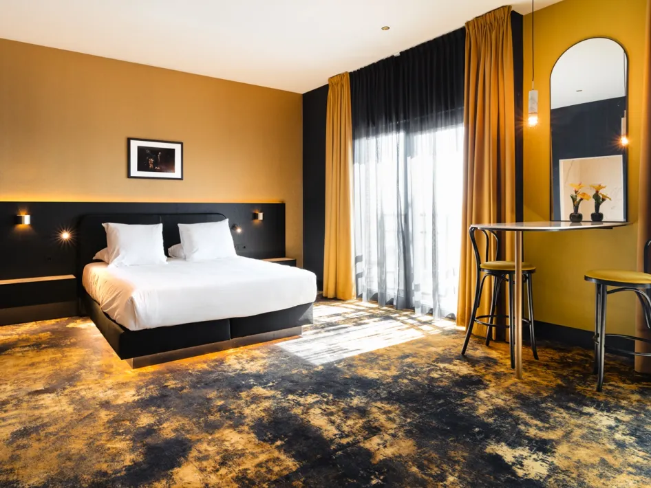 Stylish hotel room with large double bed, yellow wall and curtains and sitting area with table and chairs.