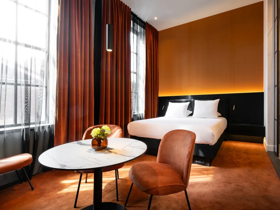 Attractive hotel room with orange curtains and wall, a double bed and a round table with chairs.