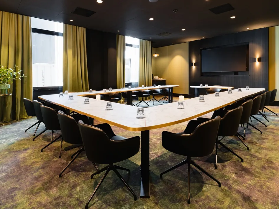 Modern meeting room with U-shaped set-up, comfortable chairs and large windows with green curtains.
