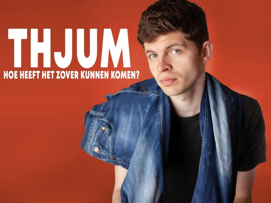 Thjum Arts, staand voor een rode achtergrond, in zwart t-shirt met een blauwe spijkerbroek hangend om zijn nek en op zijn schouder.