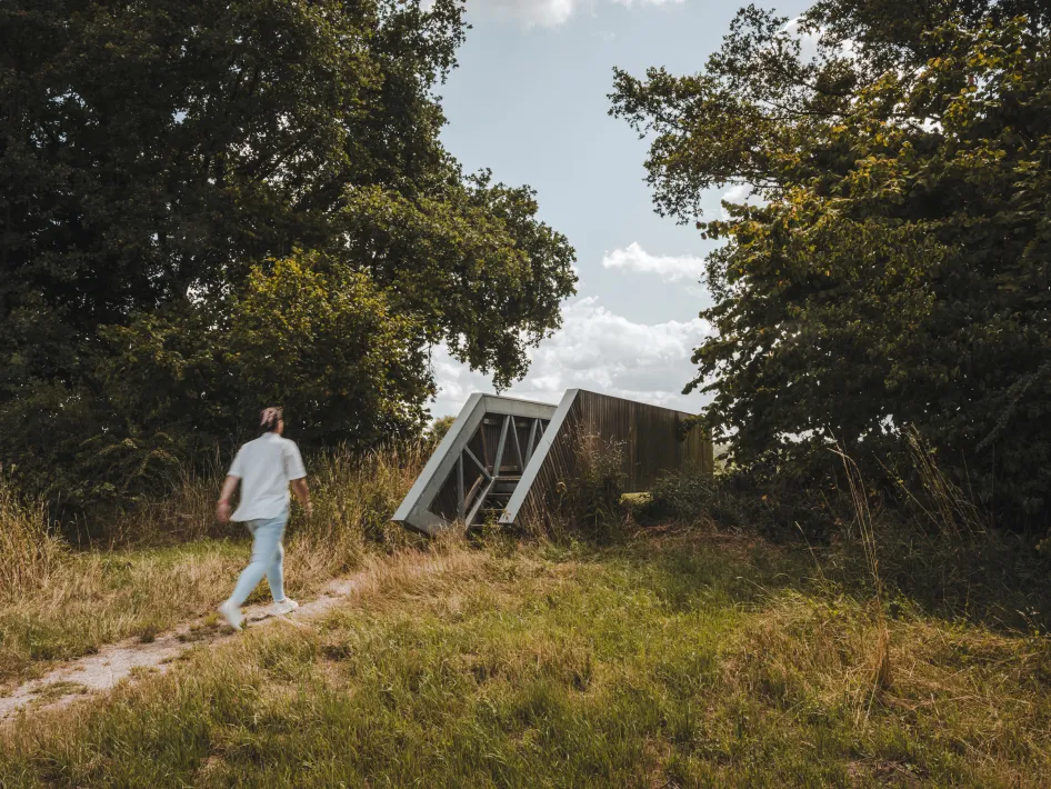 A person walks along a narrow path towards a modern wooden lookout point, partly embedded in the landscape and surrounded by tall trees and grassland under a lightly clouded sky