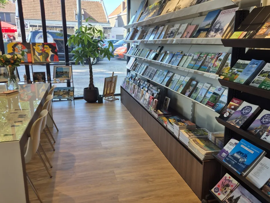 Binnenruimte van de VVV Kessel met rekken vol folders, een leestafel en grote ramen met uitzicht op de straat.