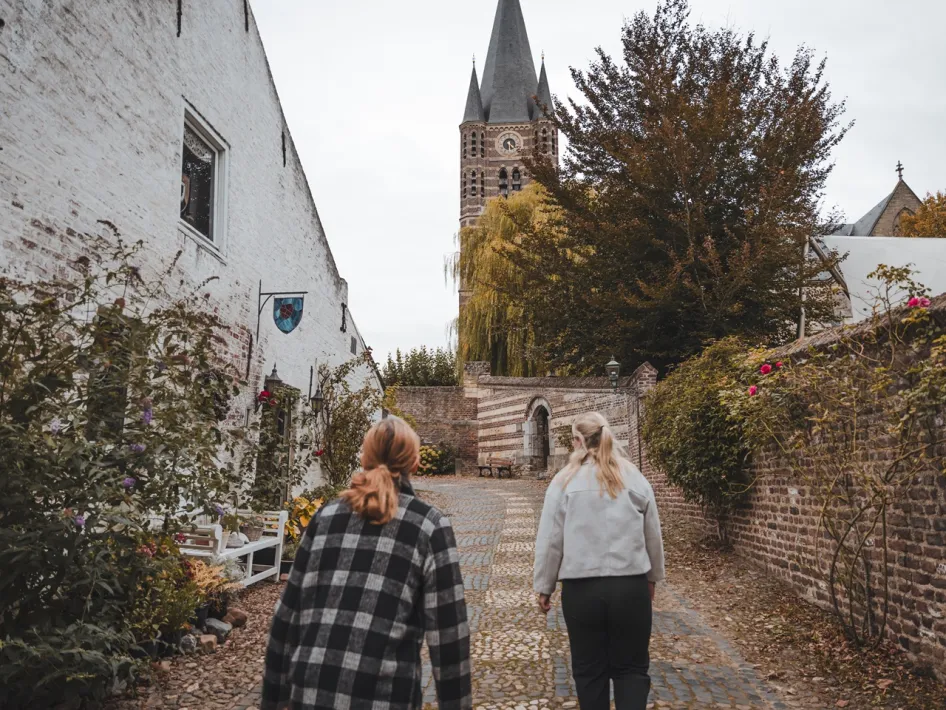 Twee vrouwen wandelen over een klinkerpad in het historische centrum van Thorn tijdens de VVV-wandeling Machtige Vrouwen van Thorn, met witte gevels, bloeiende tuinen en de kerktoren op de achtergrond.
