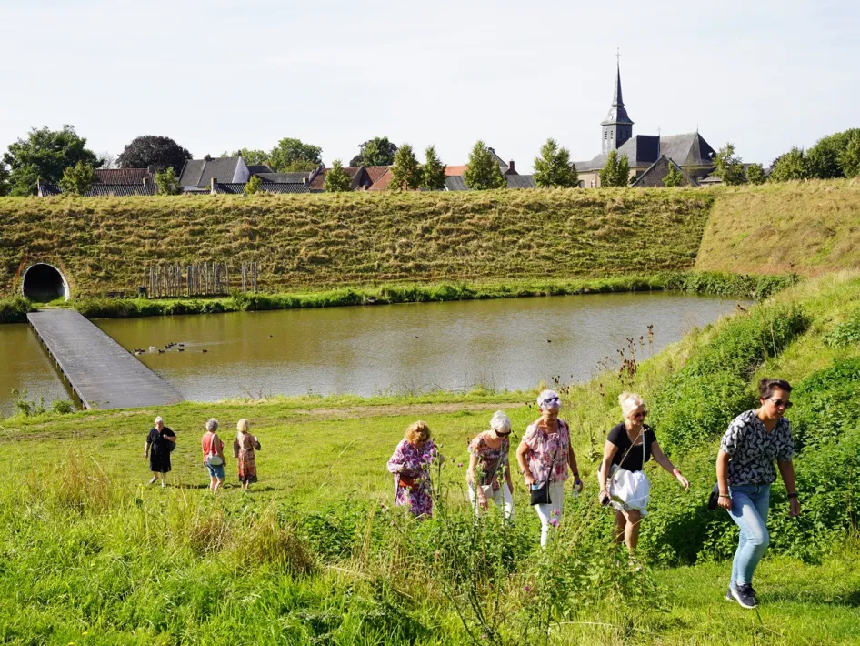 Wandelaars lopen tijdens de VVV wandeling Stevensweert, vesting aan de Maas over een groene dijk langs het water bij Stevensweert, met op de achtergrond een dorp en kerktoren.