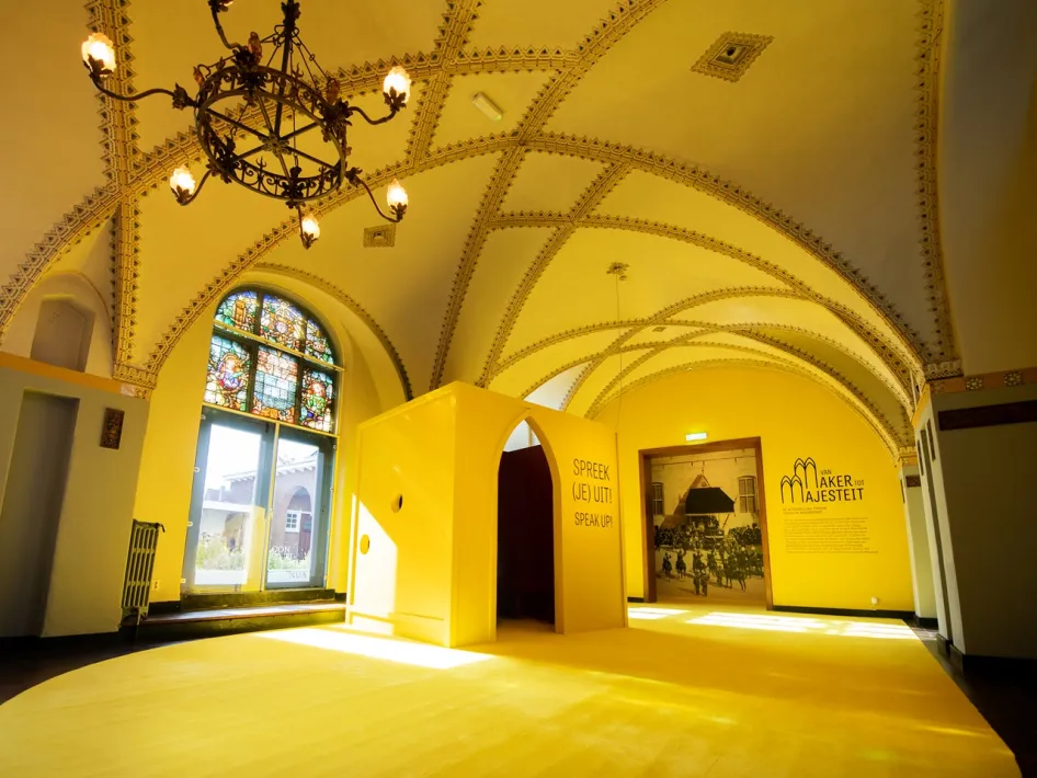 Gele tentoonstellingsruimte met de tekst 'Spreek (je) uit!' tijdens de expositie Van Maker tot Majesteit in het Cuypershuis in Roermond.