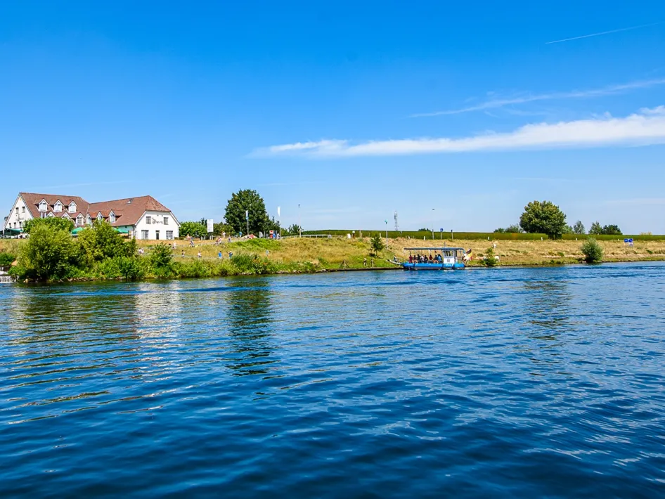 Fähre in Ohé und Laak an der Anlegestelle, im Hintergrund ein Gastronomiebetrieb an der Maas und Radfahrer am Ufer.