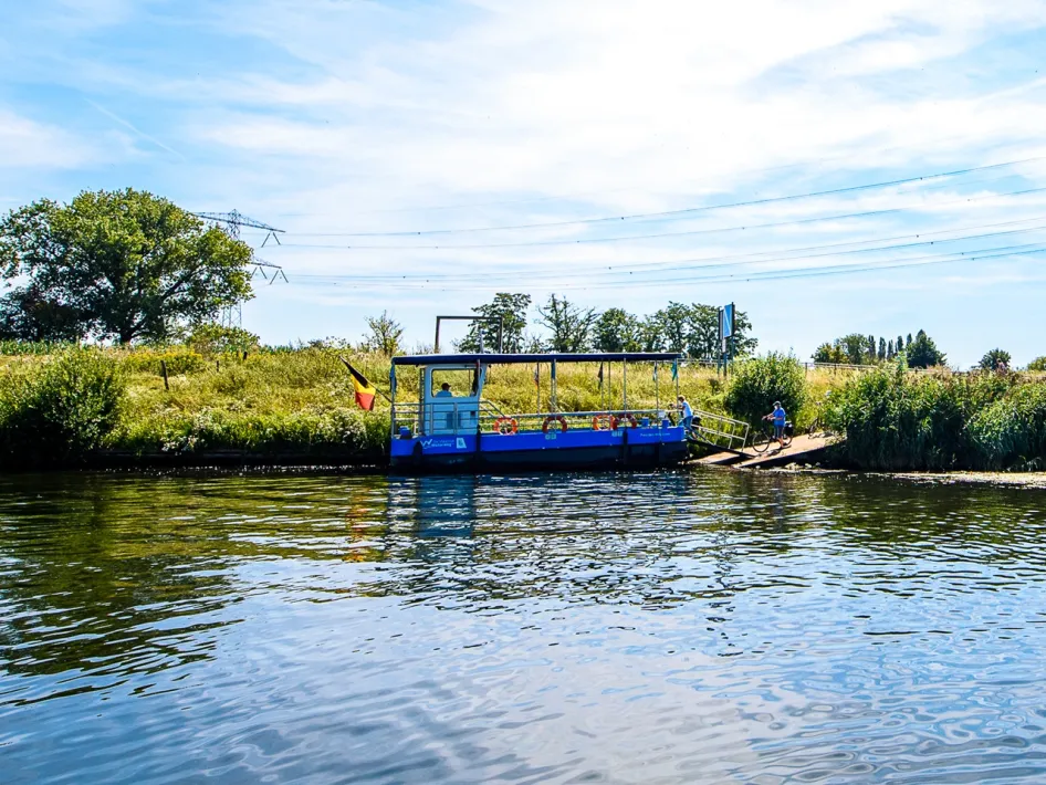 The ferry Ohé en Laak - Ophoven with Belgian flag, moors at a natural bank where a cyclist is waiting.