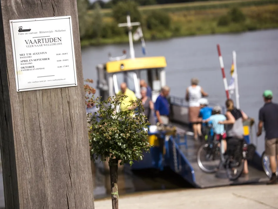 Informationstafel mit den Abfahrtszeiten der Fuß- und Fahrradfähre Blitterswijck-Wellerlooi, im Hintergrund Personen, die ihre Fahrräder auf die Fähre bringen.