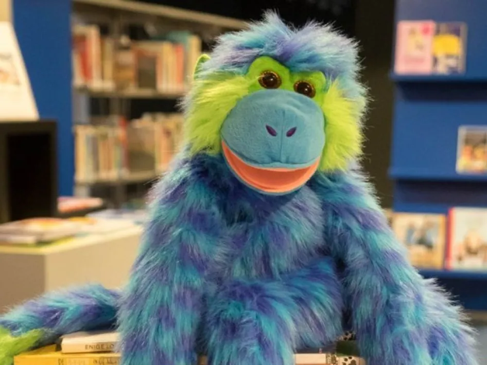 Jaap Aap, de kleurrijke handpop uit het programma Voorlezen met Jaap Aap, zit op een stapel boeken in de bibliotheek, klaar voor het voorleesmoment.