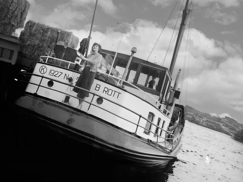 Een voortvarende vrouw hijst trots een vlag op het dek van een binnenvaartschip in de haven, met hooibalen en bedrijvigheid rondom het schip.