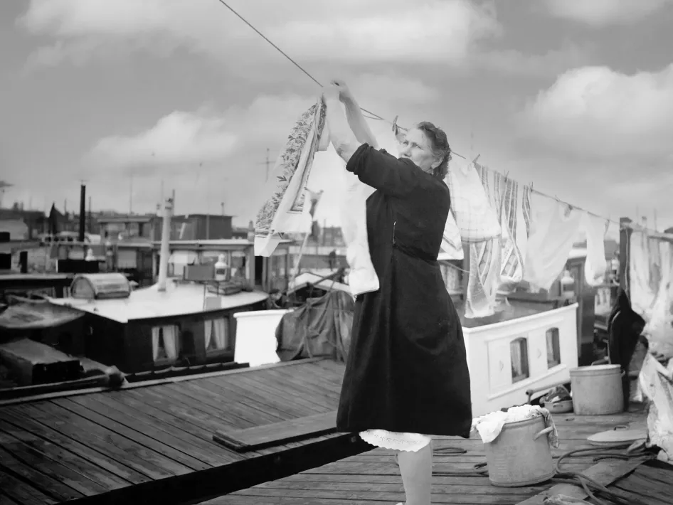 Een voortvarende vrouw hangt de was op tussen woonboten, terwijl ze op het dek staat met klompen aan – een alledaags tafereel uit het leven aan boord.