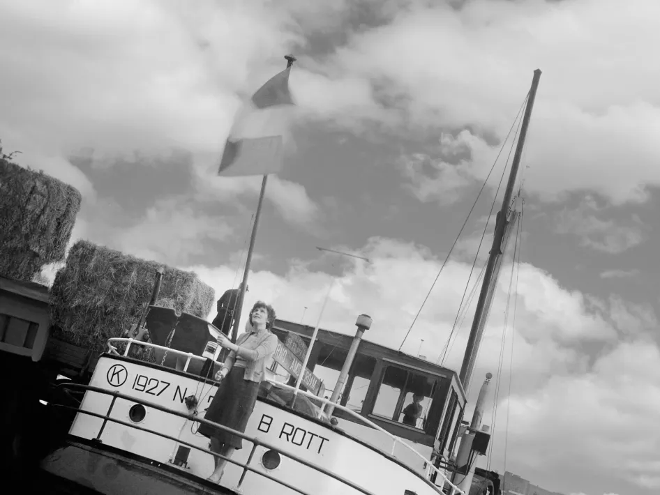 Een voortvarende vrouw hijst trots een vlag op het dek van een binnenvaartschip in de haven, met hooibalen en bedrijvigheid rondom het schip.