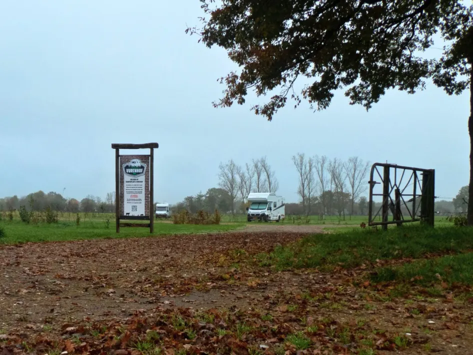 Ingang van Camperplaats Vurenhof in een groen landschap, met een informatiebord op de voorgrond en een camper geparkeerd op de achtergrond. Een open hek en herfstbladeren op het pad geven een sfeervol beeld van de locatie.