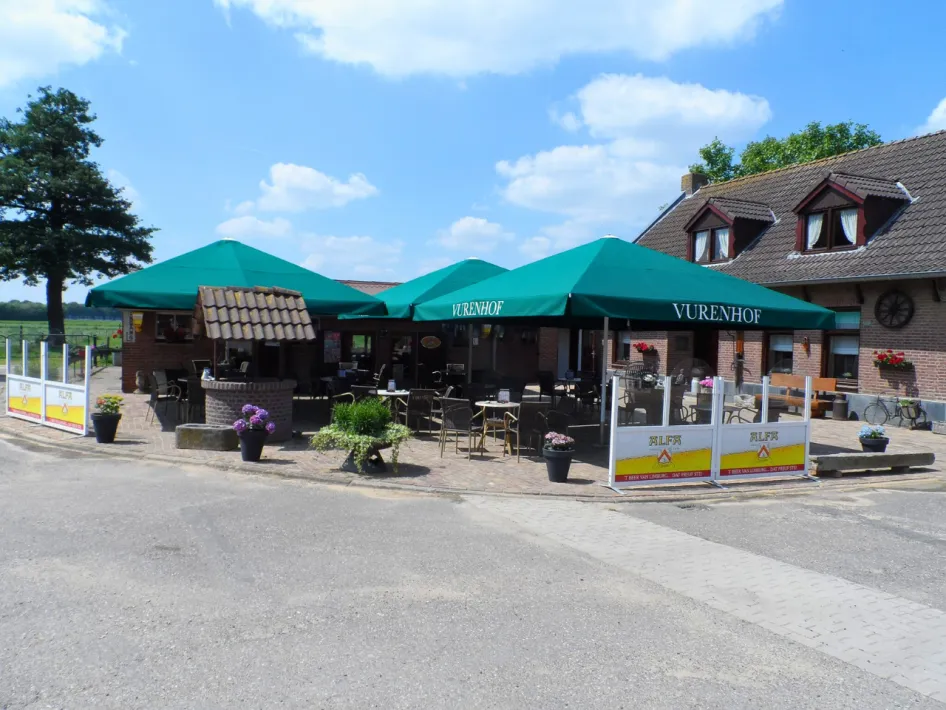 Gezellig terras van Vurenhof in Posterholt, met groene parasols, zitplaatsen en bloembakken, gelegen naast een traditioneel boerderijgebouw onder een blauwe lucht