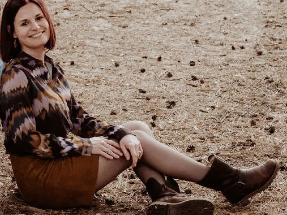 Vrouw met rood haar zit ontspannen in de natuur op een bed van dennennaalden. Ze draagt een rok en blouse met grafisch patroon en laarzen.