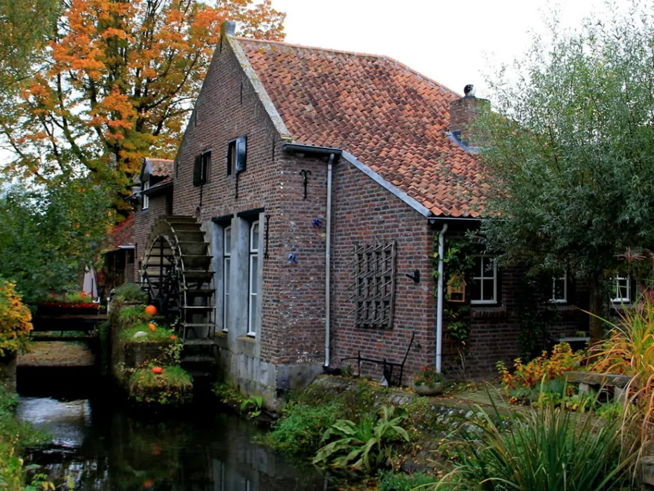 Wassermühle mit hölzernem Rad neben einem Bach bei einem Backsteinhaus, umgeben von Grün und Herbstfarben