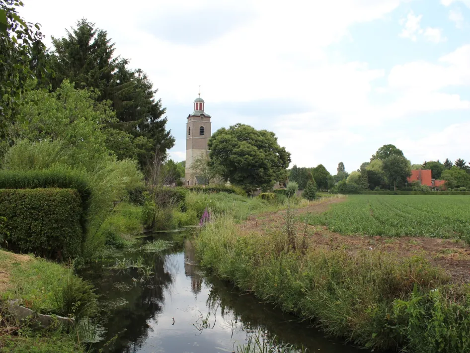 Beek die rustig door het landschap bij Neeritter stroomt, met velden, bomen en de kerktoren op de achtergrond