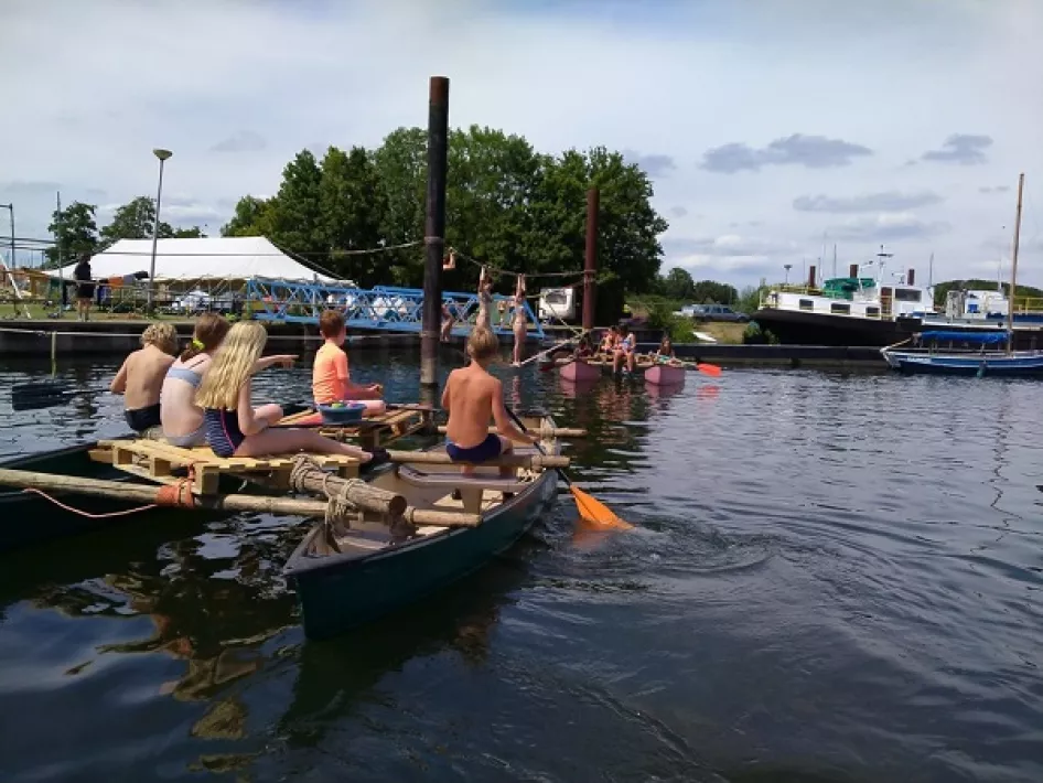 Kinderen in zwemkleding varen op zelfgebouwde vlotten en kano’s in een havengebied, met boten en een tent op de achtergrond op een zonnige dag.