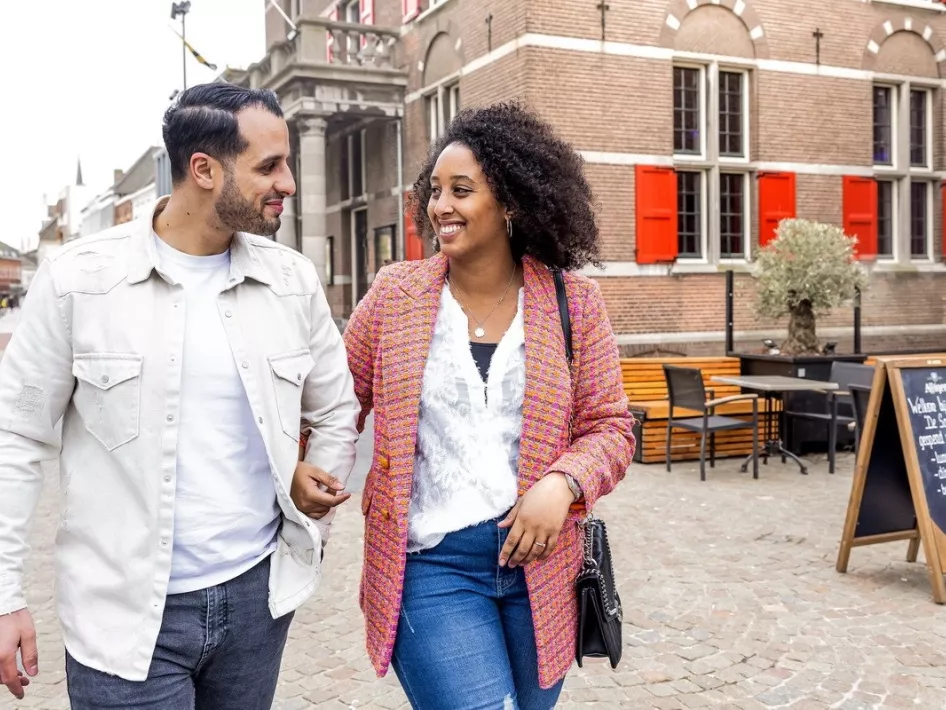 Couple walking smiling arm in arm through a historic street during the Winter Tour Weert, with a terrace in the background.