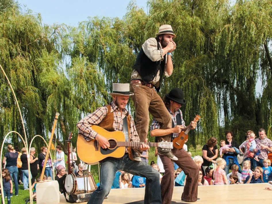 Drie muzikanten spelen op een buitenpodium met gitaar, mondharmonica en banjo, terwijl een groot publiek geniet van het optreden op een zonnige dag.