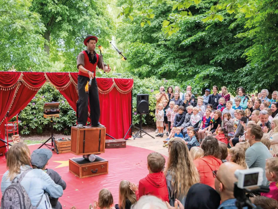 Een artiest balanceert op een kist terwijl hij jongleert voor een groot publiek tijdens een openluchtvoorstelling met een rood theaterdecor.