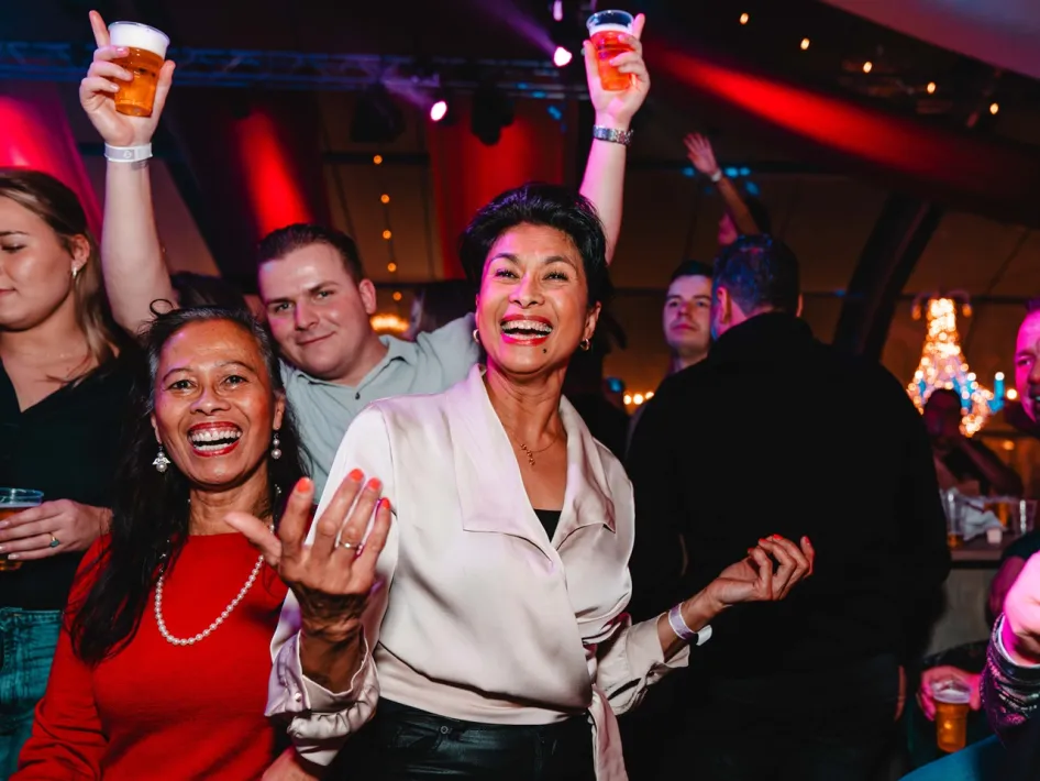 Twee vrouwen zijn aan het dansen terwijl op de achtergrond een man zijn armen in de lucht gooit en die twee biertjes vastheeft.