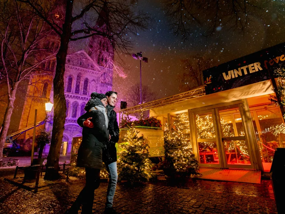 Ein Paar spaziert gemütlich durch den Schnee an einem beleuchteten Winterpavillon auf dem Munsterplein in Roermond vorbei.