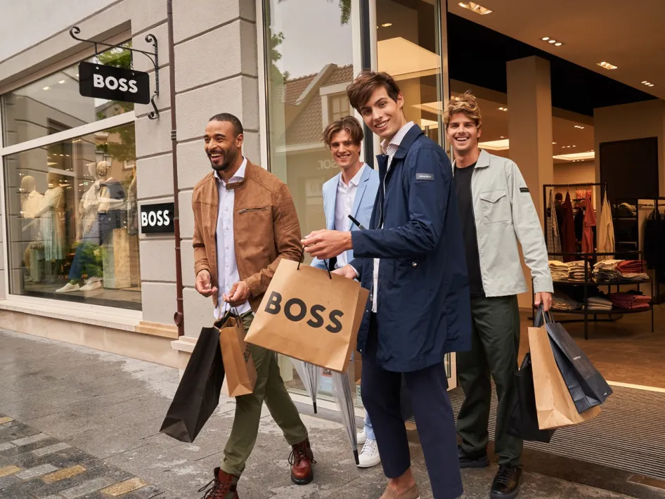 Vier Männer mit Einkaufstaschen verlassen lachend das BOSS-Geschäft während der Wintersale im Designer Outlet Roermond.