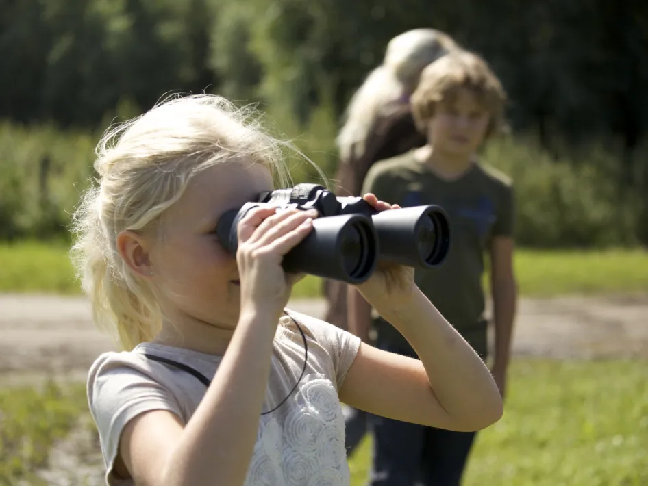 Een blond meisje kijkt door een verrekijker tijdens een natuurwandeling. Op de achtergrond lopen een jongen en een volwassene over een bospad.