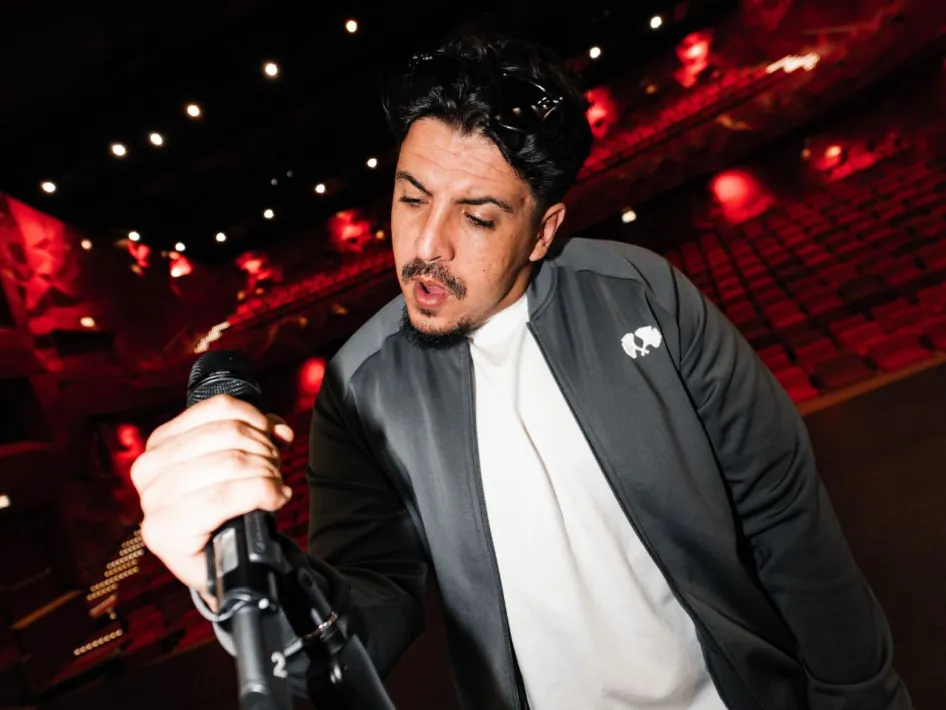 Rapper en acteur ICE (Khalid Alterch) houdt een microfoon vast in een theaterzaal met rode verlichting.