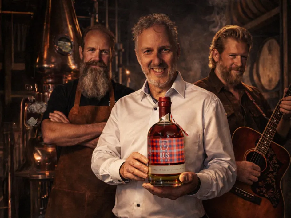 Drie mannen in een whiskystokerij, met op de voorgrond een man die een fles BUS Whisky vasthoudt en rechts een muzikant met gitaar.
