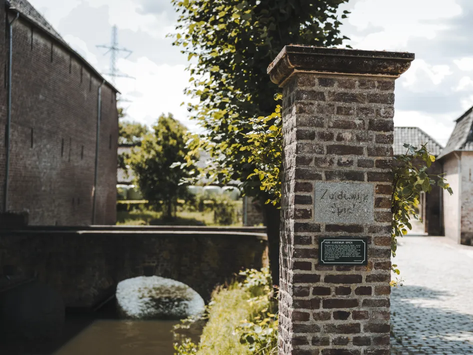 Eine gemauerte Säule mit der Inschrift „Zuidewijk Spick“ markiert den Eingang zu einem historischen Landgut. Im Hintergrund sind eine alte Steinbrücke über einen Wassergraben und Backsteingebäude unter einem teilweise bewölkten Himmel zu sehen.