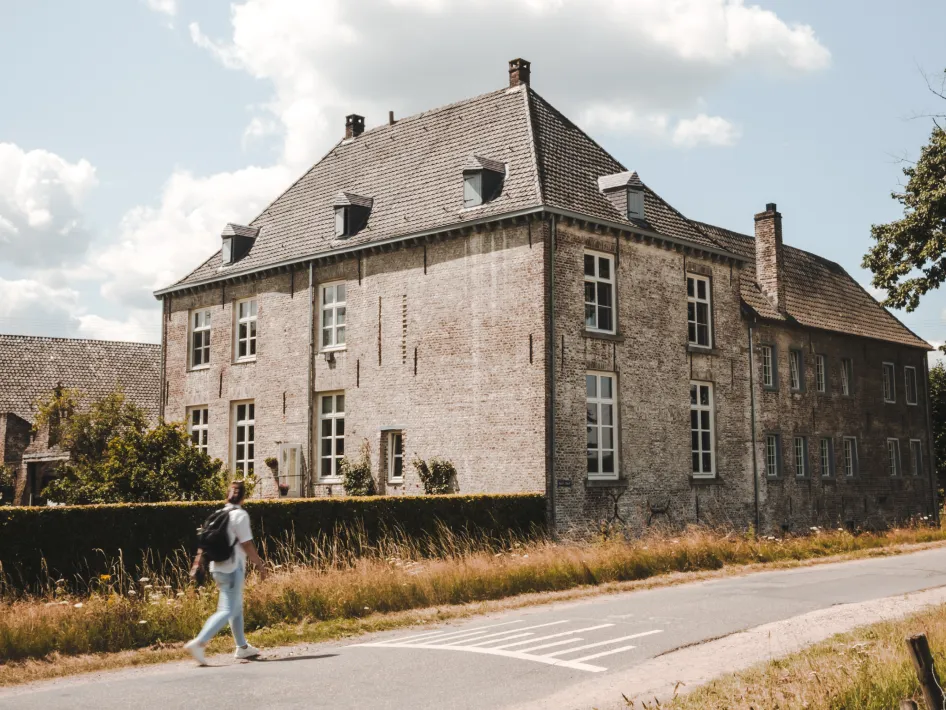 Eine Person geht an einem großen, alten Herrenhaus aus Ziegelstein mit hohen Fenstern und einem Satteldach vorbei. Das Gebäude liegt an einer ruhigen Straße und ist teilweise von einer Hecke und Blumen umgeben.