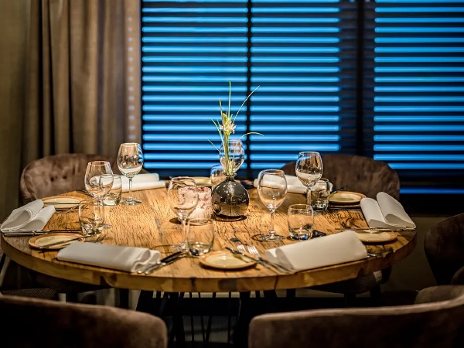 Rond houten restauranttafel sfeervol gedekt met wijnglazen, servetten en een vaasje met bloemen.