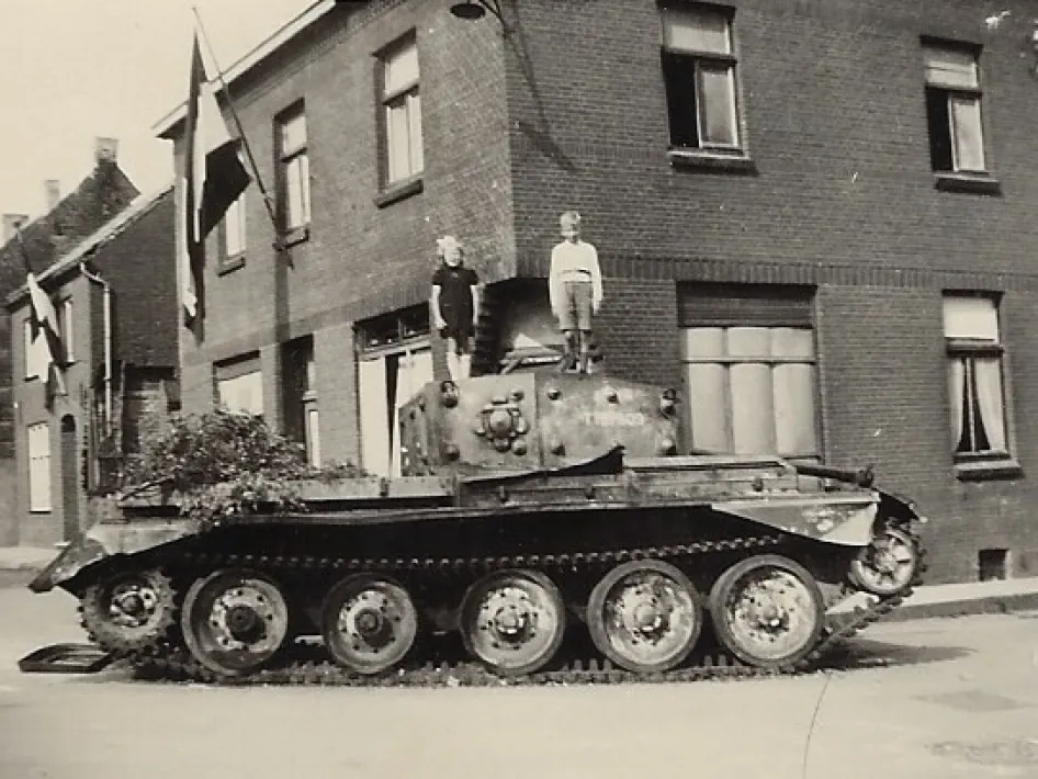 Zwart-witfoto van een beschadigde tank op een straathoek in een Nederlands dorp, vermoedelijk uit de Tweede Wereldoorlog. Twee kinderen staan bovenop de tank. Aan de omliggende huizen hangen vlaggen, wat wijst op een bevrijdingsmoment.
