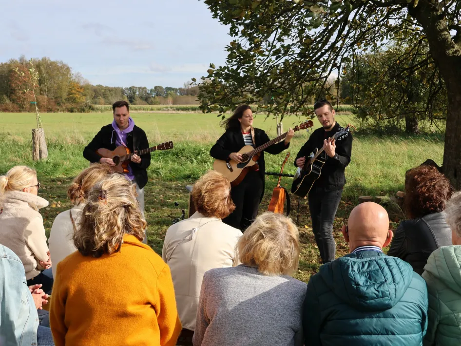 Drie muzikanten spelen gitaar voor een zittend publiek in een open veld, onder een boom, op een zonnige herfstdag.