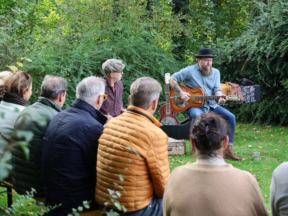 Een kleine groep mensen luistert aandachtig naar een man met een baard en hoed die gitaar speelt tijdens een intiem buitenconcert in een groene bosrijke omgeving.
