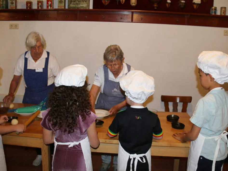 Kinderen met bakkersmutsen en schorten die samen deeg maken tijdens een bakmiddag in het museum, begeleid door vrijwilligers aan tafel.