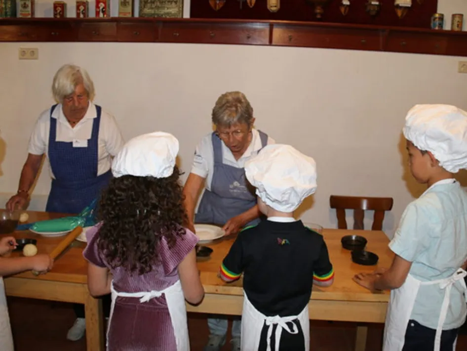Kinderen bakken samen koekjes tijdens een bakmiddag in het museum, met deeg, deegrollen en bakkersspullen op tafel.