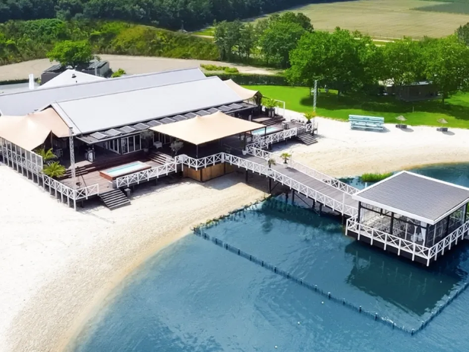 Luchtfoto van Beachclub Degreez in Panheel, waar het weddingevent plaatsvindt, gelegen aan het water met een zandstrand, houten terrassen en een steiger die uitkijkt over het meer, omringd door groen landschap.