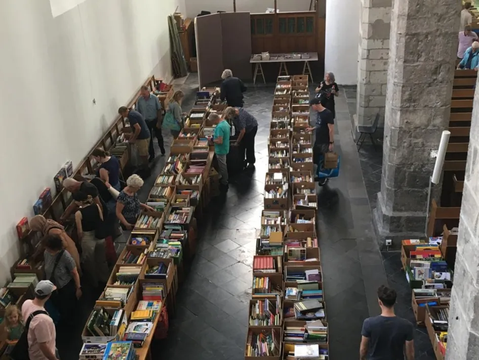 Bezoekers snuffelen tussen lange tafels vol boeken tijdens een drukbezochte boekenmarkt Minderbroederskerk  in een kerk, kom zelf ook rondkijken en ontdek mooie vondsten