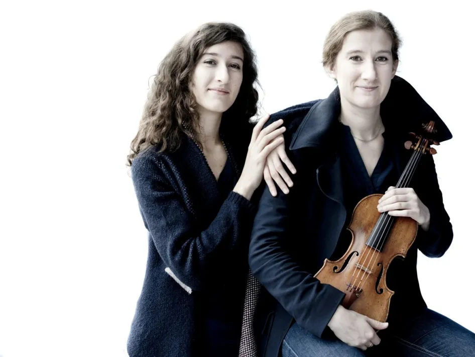 Twee vrouwelijke muzikanten poseren samen, één met een viool in de hand, tegen een lichte achtergrond.