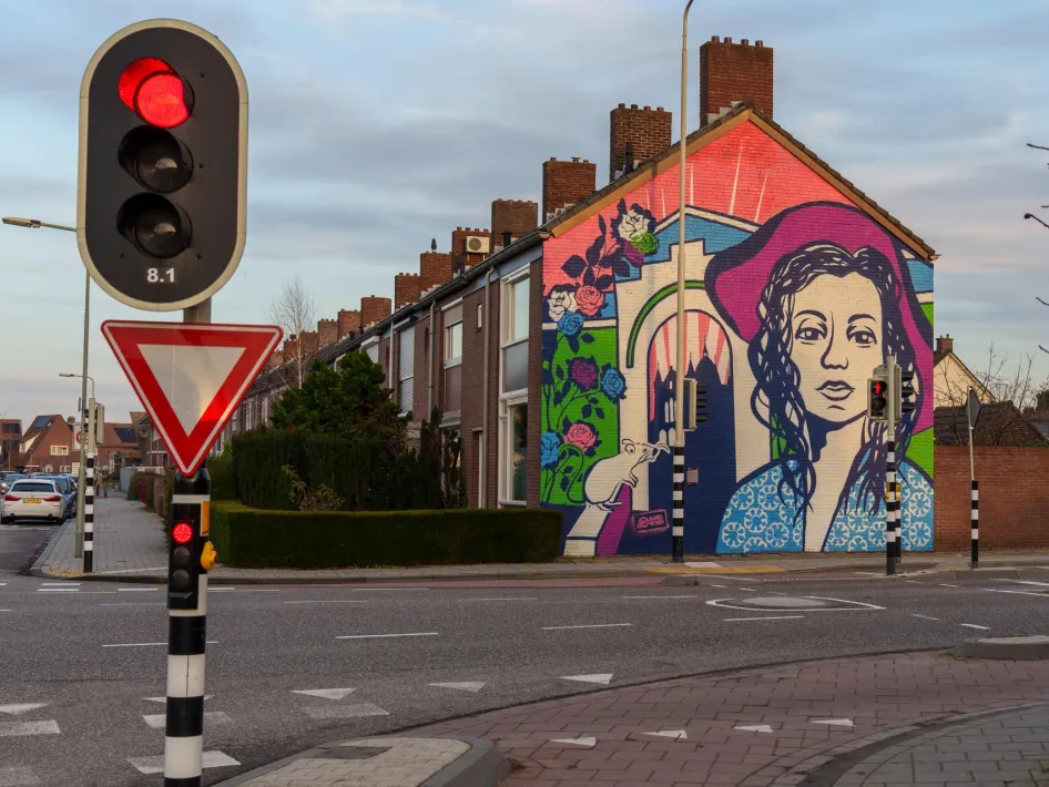 Muurschildering van een vrouwenportret in felle kleuren op de kopgevel van een rijtjeshuis aan een kruispunt met verkeerslichten.